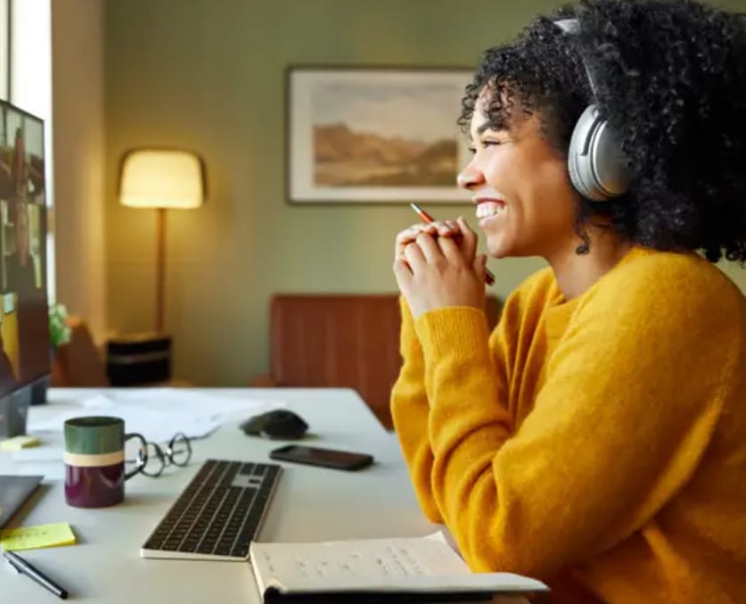 A woman with curly black hair and wearing a yellow sweater is sitting at a desk with a computer. She's wearing silver headphones and holding a pen to her mouth as if deep in thought or listening intently. The desk has a keyboard, mouse, mug, glasses, and papers. A lamp and a framed picture are in the background.