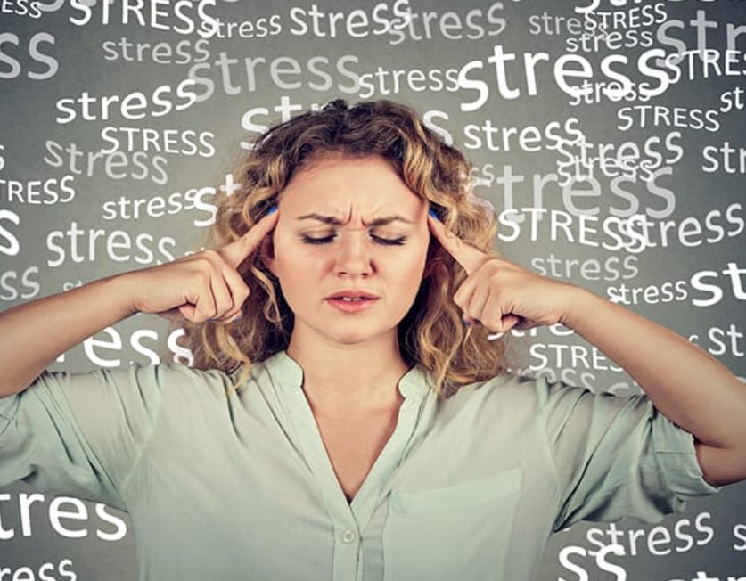 The image shows a woman with blonde hair and a light-colored shirt, holding her hands to her temples, conveying stress or frustration. The grey background has the word "stress" written repeatedly, emphasizing her anxious expression and tense posture.