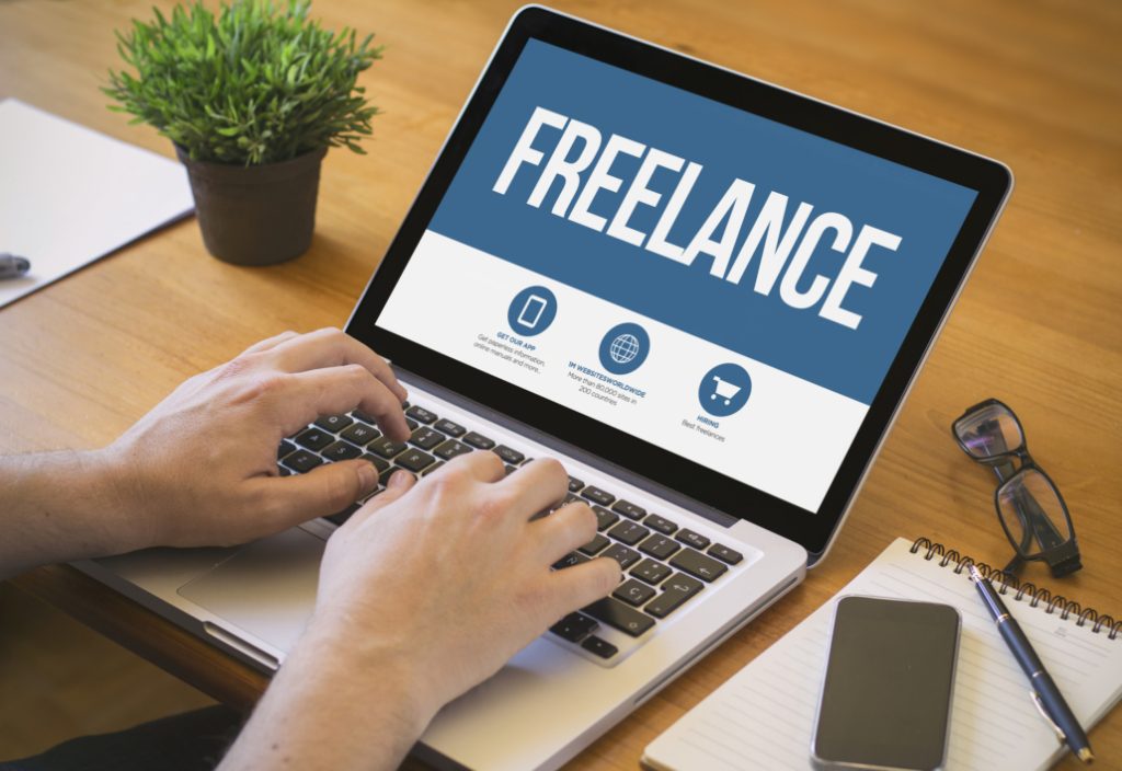 The image shows a person working on a laptop with the word "FREELANCE" prominently displayed on the screen.

- The laptop is on a wooden desk with a person's hands typing on the keyboard.
- The screen has a blue and white design with "FREELANCE" in large white letters.
- Below the title are three icons with text:
    - "GET OUR APP" with a phone icon
    - "WORK WORLDWIDE" with a globe icon
    - "HIRING" with a shopping cart icon
- Other items on the desk include:
    - A small potted plant (top left)
    - A notebook and pen (bottom right)
    - A smartphone (next to the notebook)
    - A pair of glasses (next to the smartphone)