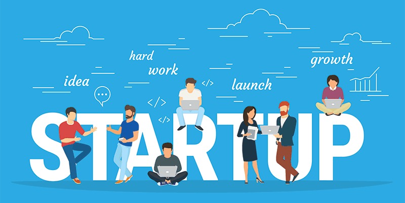 The image depicts a vibrant illustration of the word "STARTUP" in large white letters against a bright blue background.

Seven people are positioned around the letters, each engaged with laptops in various poses like sitting, standing, or crouching. Surrounding the word are keywords like "idea", "hard work", "launch", and "growth" in white cursive text, accompanied by simple graphics such as clouds, lines, a speech bubble, code symbols, and a bar graph. The overall vibe is energetic and suggests teamwork, innovation, and progress in a startup environment.