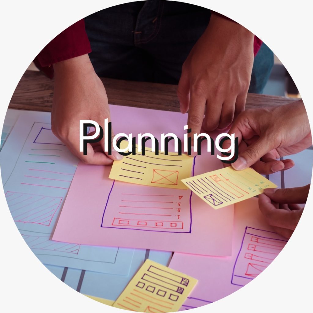 The image shows two people’s hands working at a wooden table covered with colorful paper sketches of website or app wireframes (pink and blue sheets with UI layouts) and yellow sticky notes that have mock email icons and content blocks. The word “Planning” is overlaid in large white-and-black text across the center, indicating the scene depicts a collaborative planning or design brainstorming session for a digital interface.