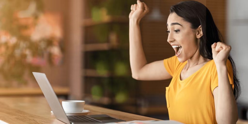 This image shows a young woman celebrating in front of her laptop. She is wearing a yellow t-shirt and has her fists raised in excitement, her mouth open in a joyful expression. On the table in front of her is a cup of coffee and the laptop she is looking at. The background is softly blurred, suggesting a cozy home or café environment.

This image could visually represent the excitement or satisfaction a job seeker feels after achieving a goal, such as learning something new, completing a course, or landing an interview or job. It pairs well with the theme of maintaining a learning journal, showing how consistent tracking and reflection can lead to positive outcomes.