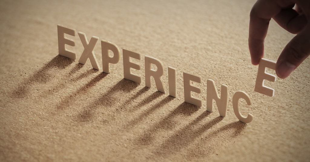 A hand is placing the letter “E” to complete the word “EXPERIENCE,” which is formed with standing 3D letters on a textured, sandy-colored surface. The letters cast long shadows, giving a sense of depth and perspective. The image symbolizes building, completing, or gaining experience step by step.
