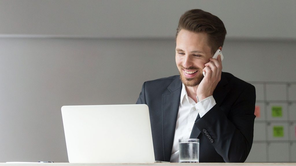 A smiling professional man wearing a black suit and white shirt, sitting at a desk while talking on a smartphone and looking at a laptop screen, suggesting a phone or virtual interview setup.