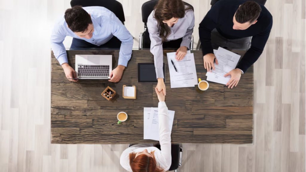 Top view of a professional job interview scene where a candidate is shaking hands with an interviewer across a table, while other interviewers review documents and work on a laptop, representing hiring and recruitment discussions.