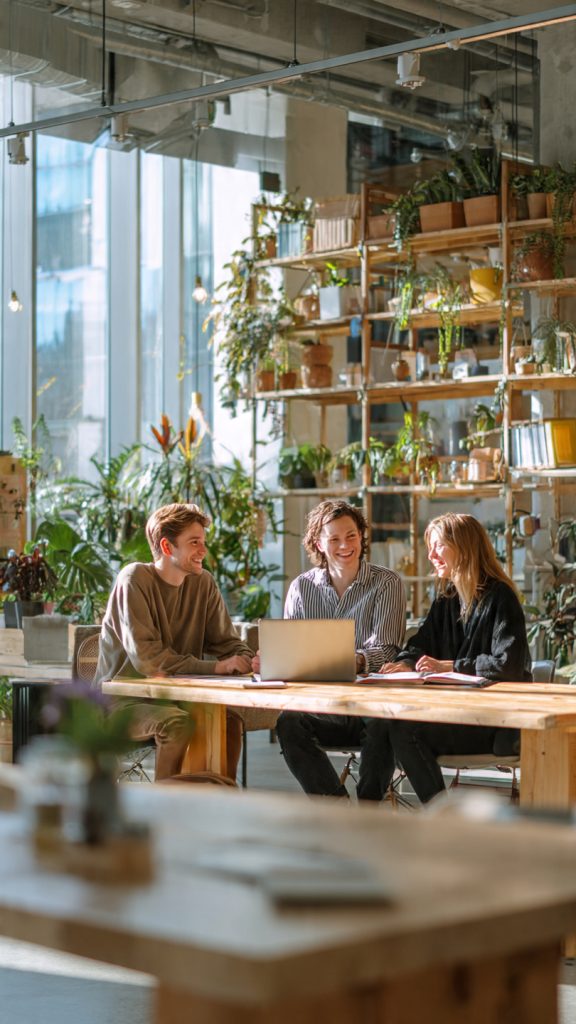 Design Onboarding
create image Three professionals sitting around a wooden table in a modern, sunlit office, smiling and engaged in conversation with a laptop open between them, surrounded by plants, shelves, and large windows in a bright, collaborative workspace.